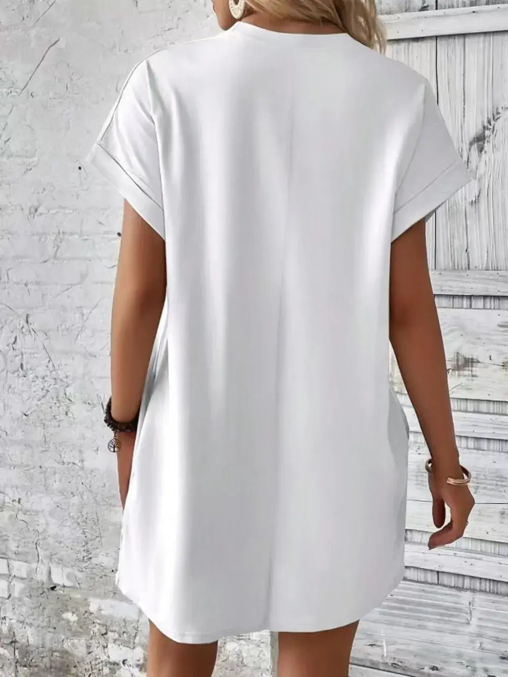 Back view of a woman wearing a white pocketed round neck short sleeve dress against a textured wall.