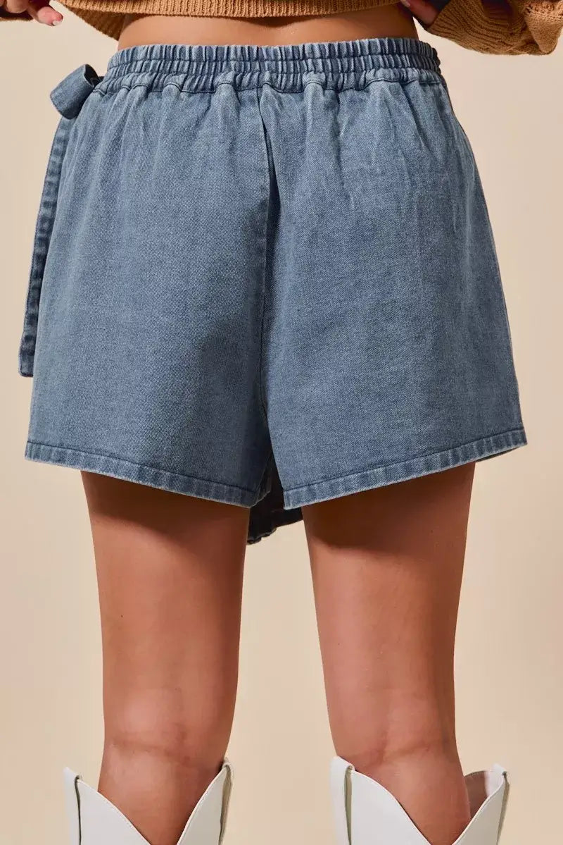 Back view of a woman's denim skort featuring a wrap design and braided trim, showcasing its trendy and chic style.