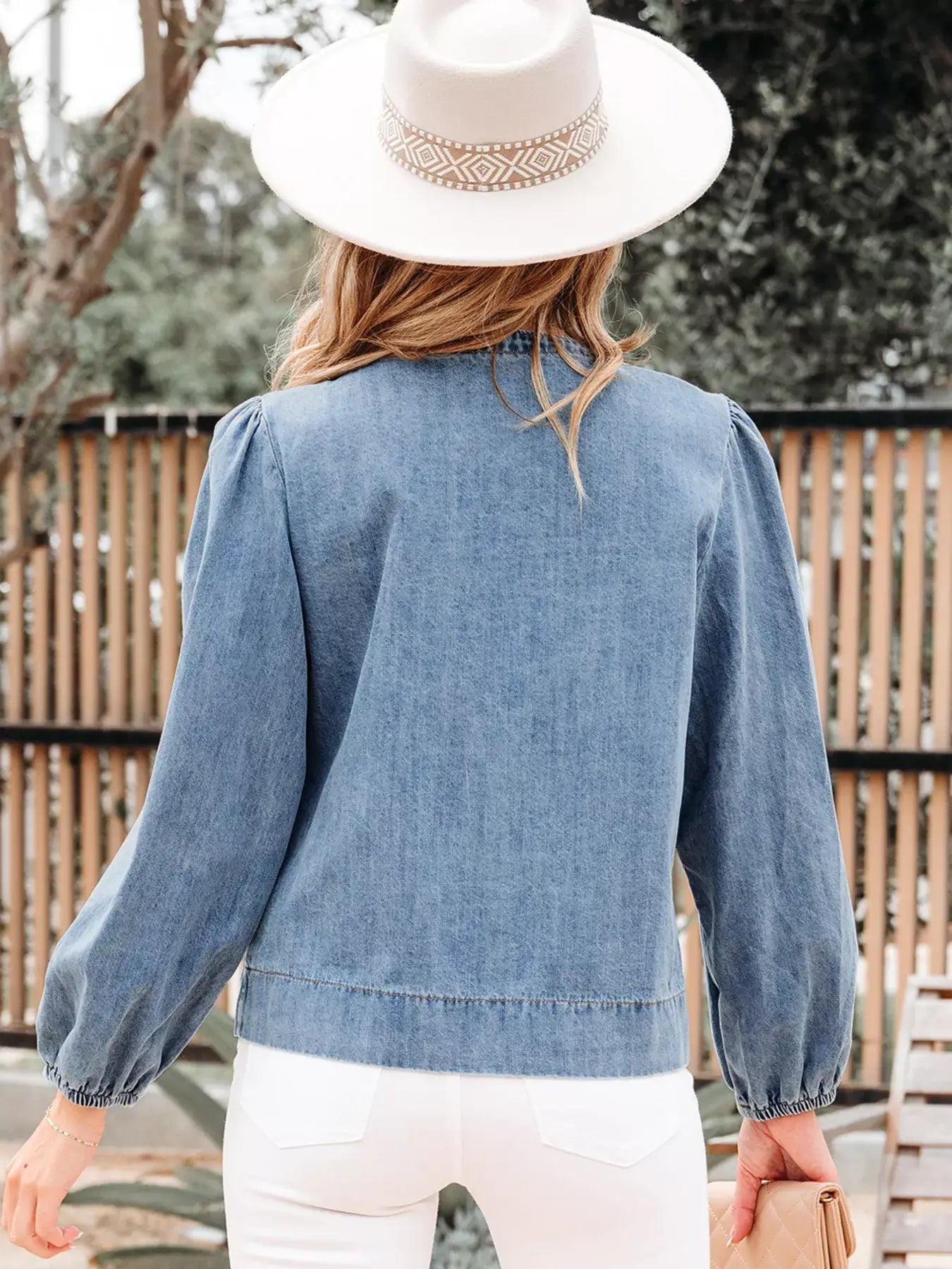 Back view of a woman wearing a tied front puff sleeve denim top, paired with white pants and a stylish hat.
