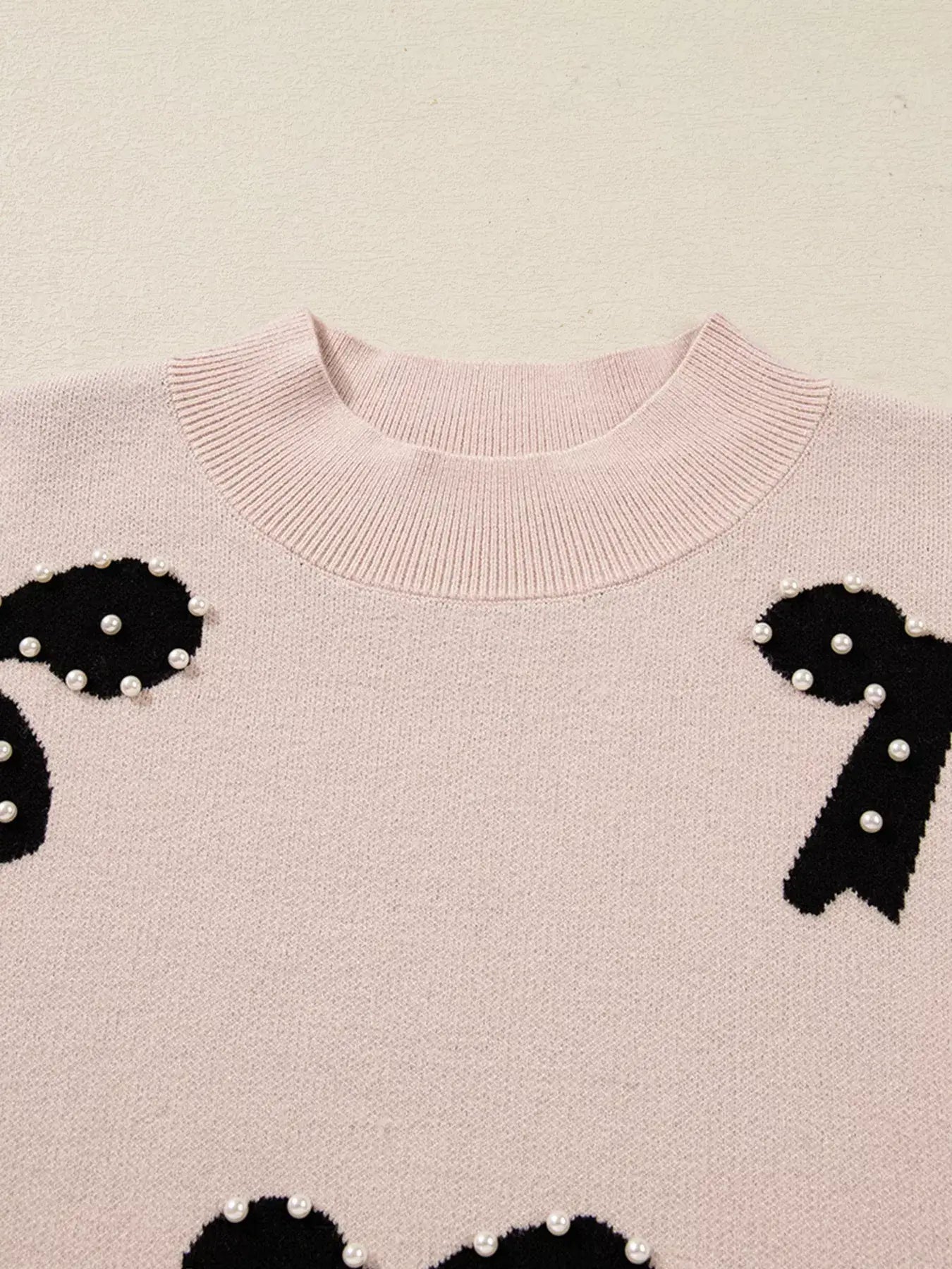 Pearl embellished bow pattern loose sweater featuring a soft pink knit with black bow accents and pearl embellishments.
