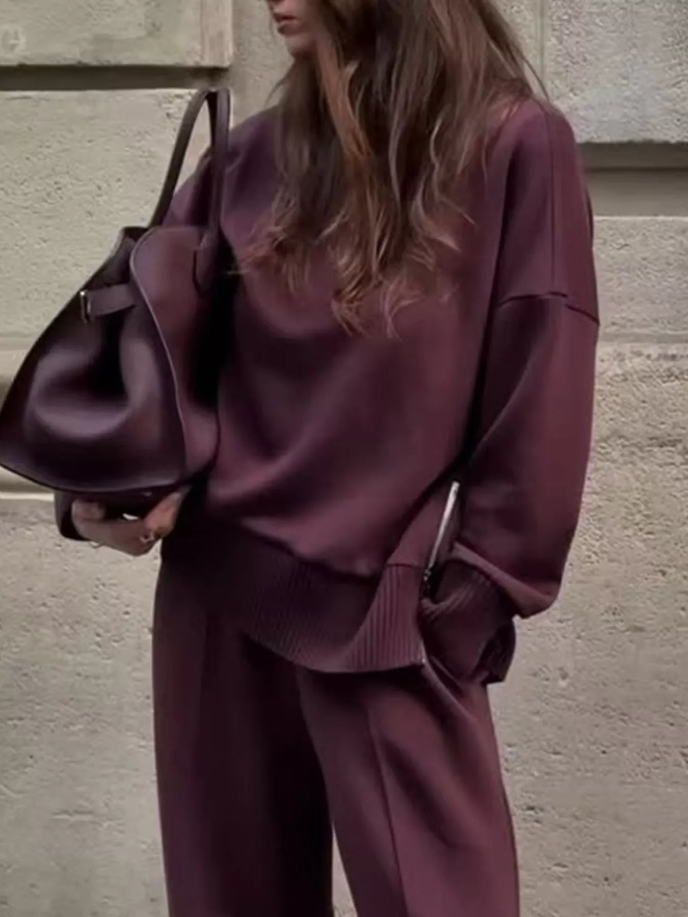 Model wearing Turtleneck Side Zipper Sweatshirt Pants Set in burgundy, holding a bag against a textured wall.