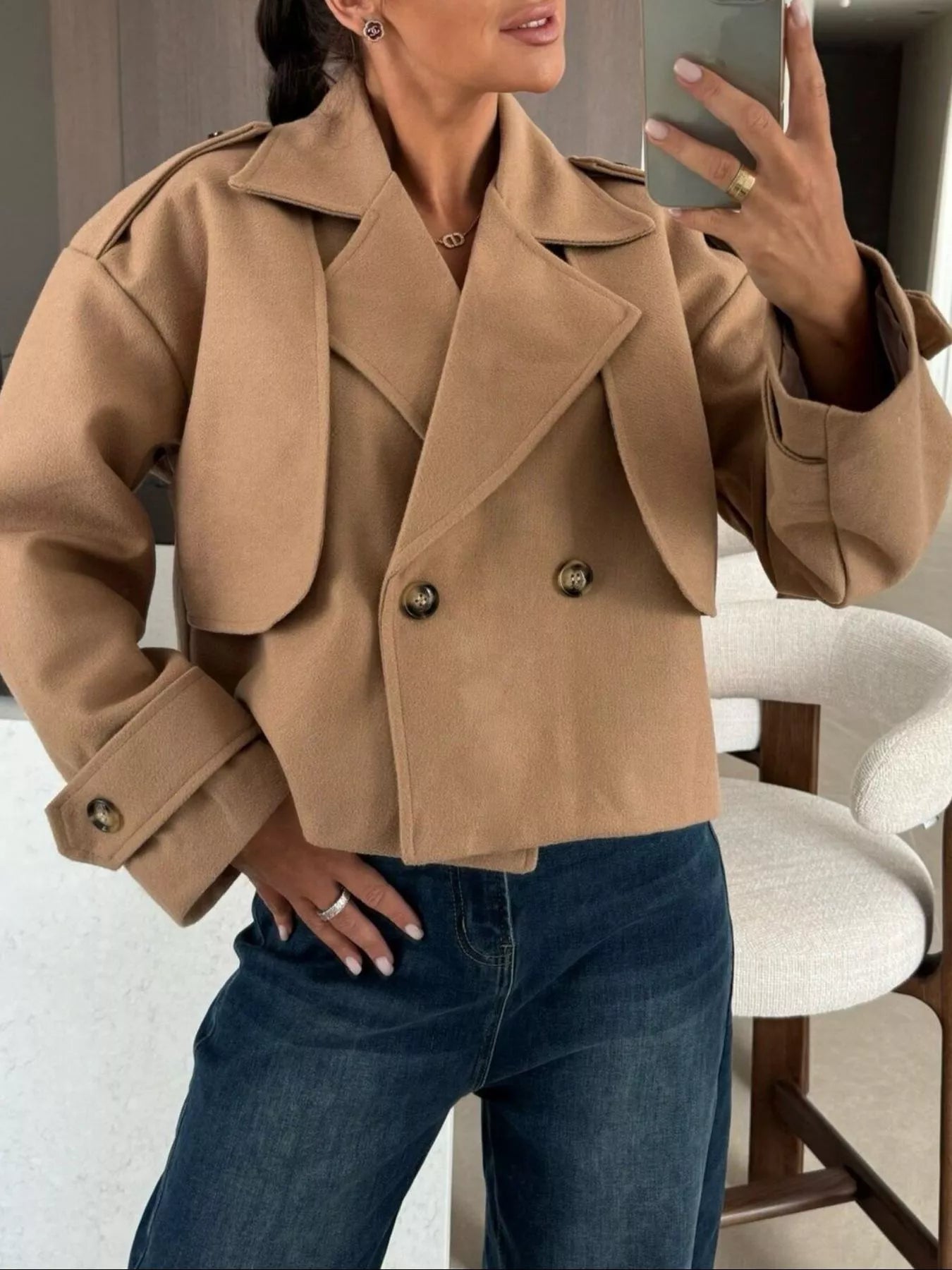 Double-Breasted Cropped Outerwear in tan color styled with jeans and a hand on the mirror, showcasing a modern look.