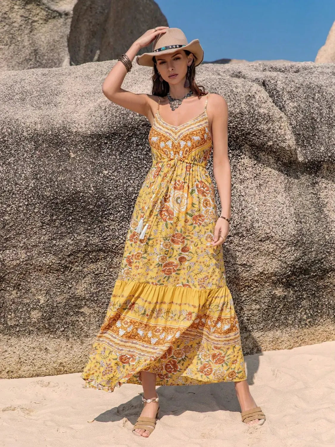 Model wearing a Tassel Printed Tiered Cami Dress in yellow with floral print, on the beach with a sun hat and charm accessories.