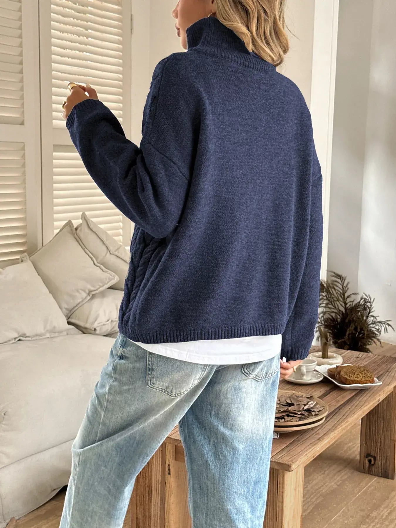 Back view of a woman in a zip collar cable-knit sweater, showcasing the chunky knit design in a cozy indoor setting.