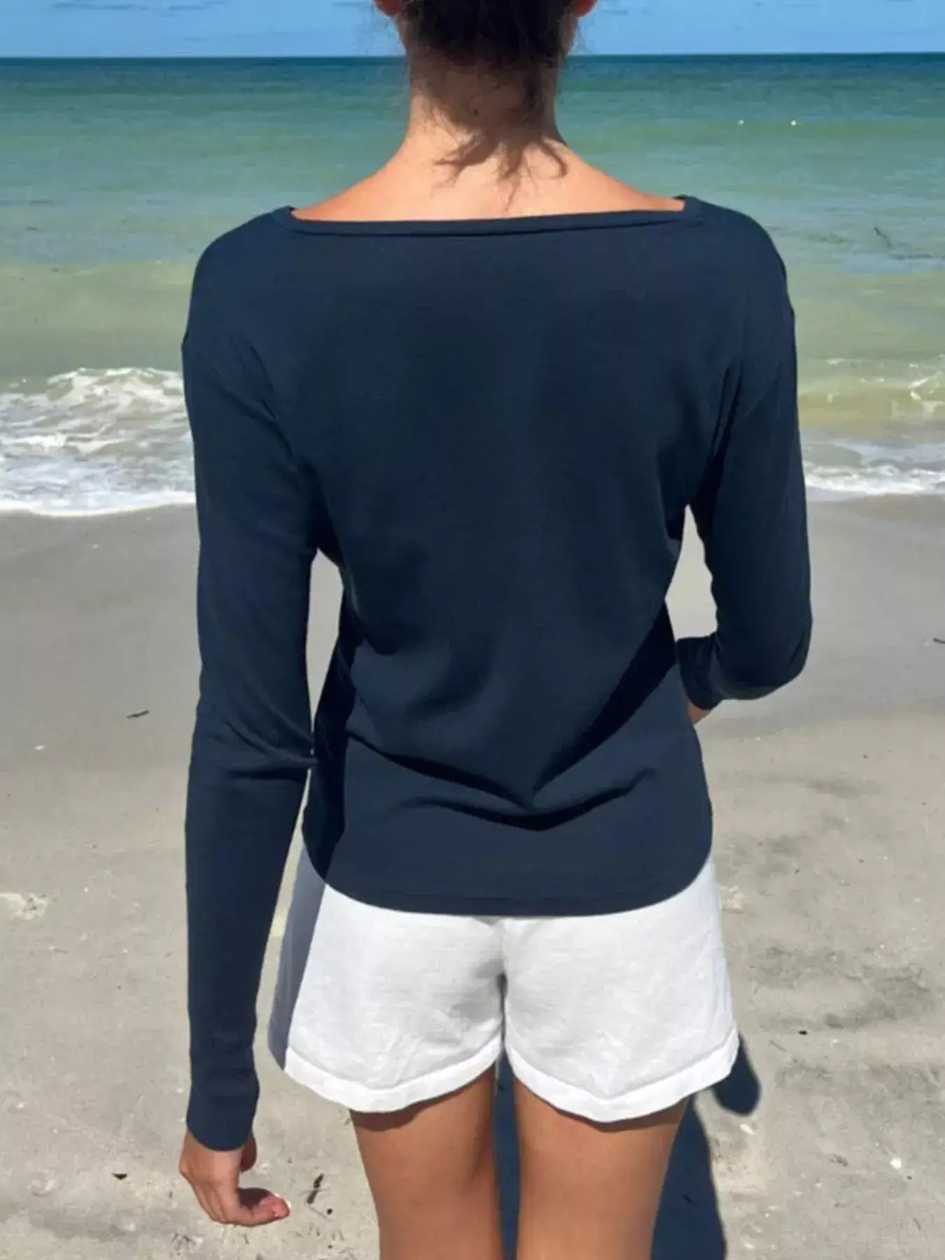 Back view of a woman wearing a navy long sleeve top and white shorts on a beach.