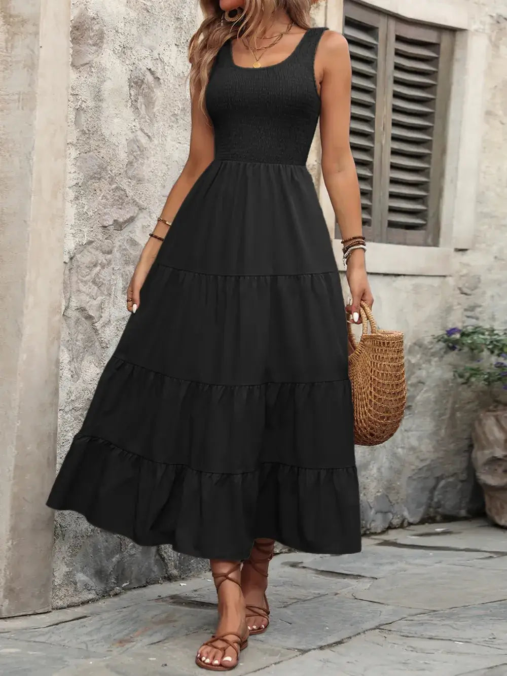 Black sleeveless smocked scoop neck maxi dress with tiered skirt, stylishly paired with a woven bag.
