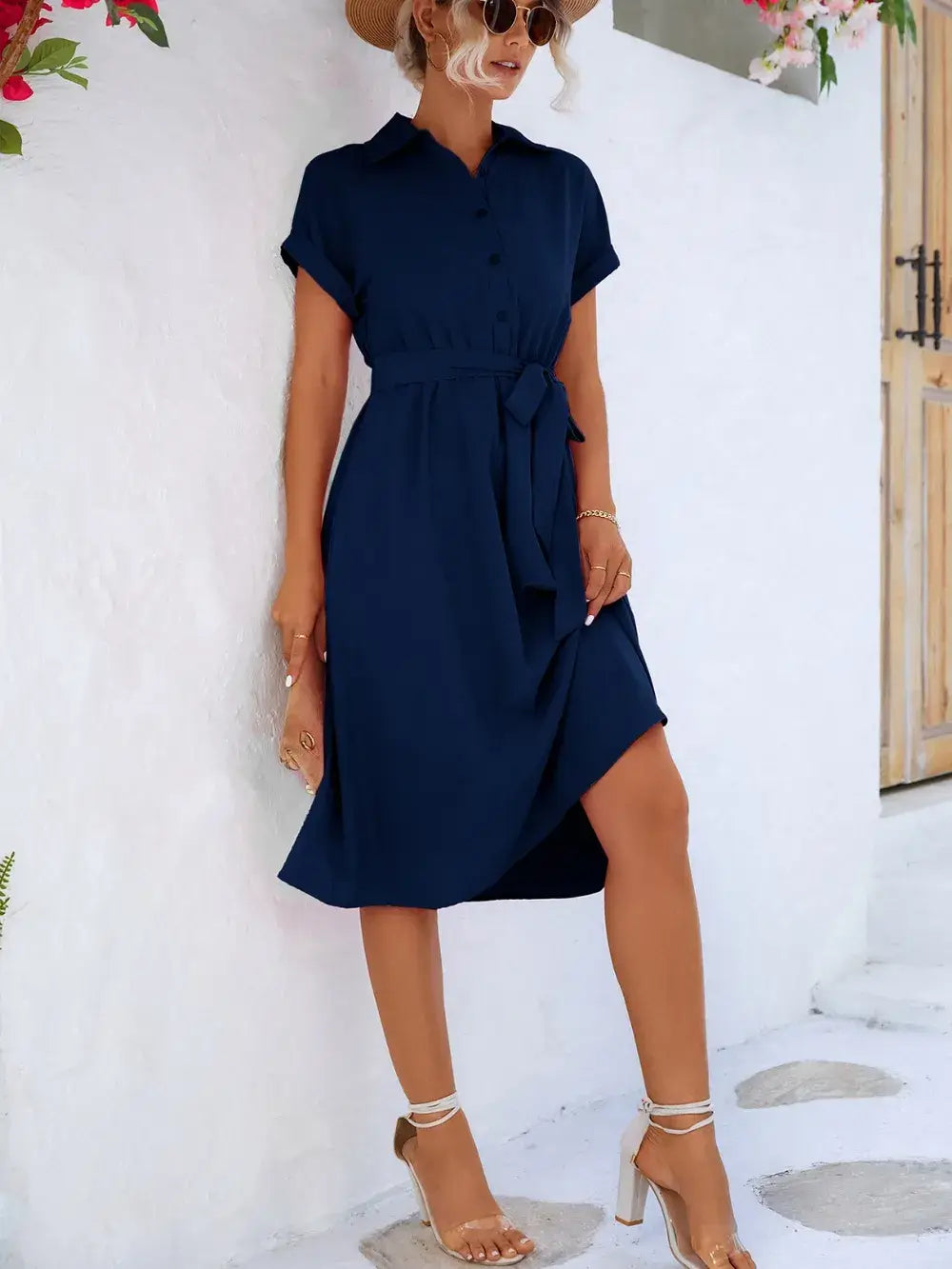 Model wearing a navy buttoned tie waist short sleeve dress against a white wall, showcasing a casual summer style.