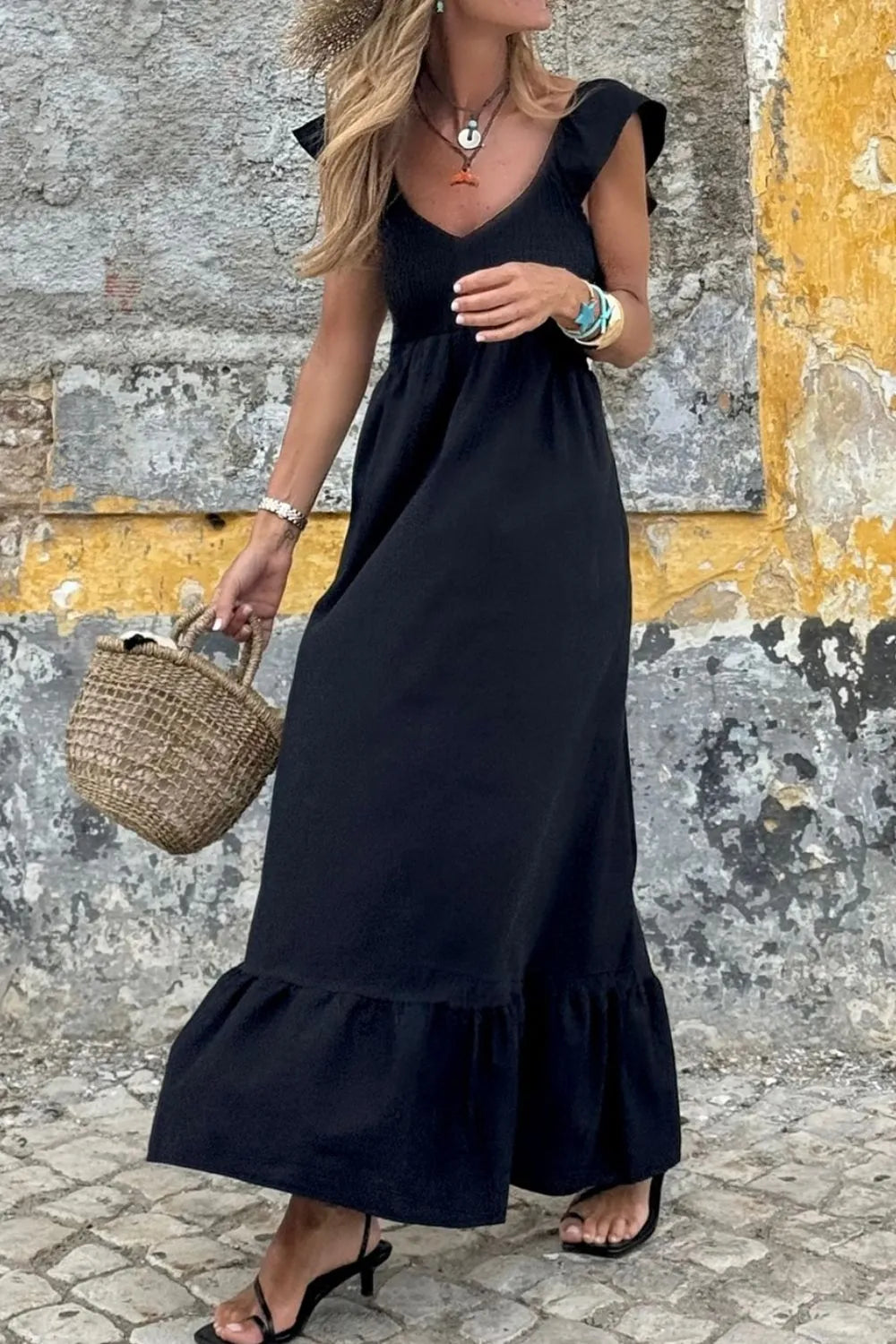 Model wearing a V Neck Ruffled Cap Sleeve Smocked Maxi Dress in black against a textured wall with a basket.