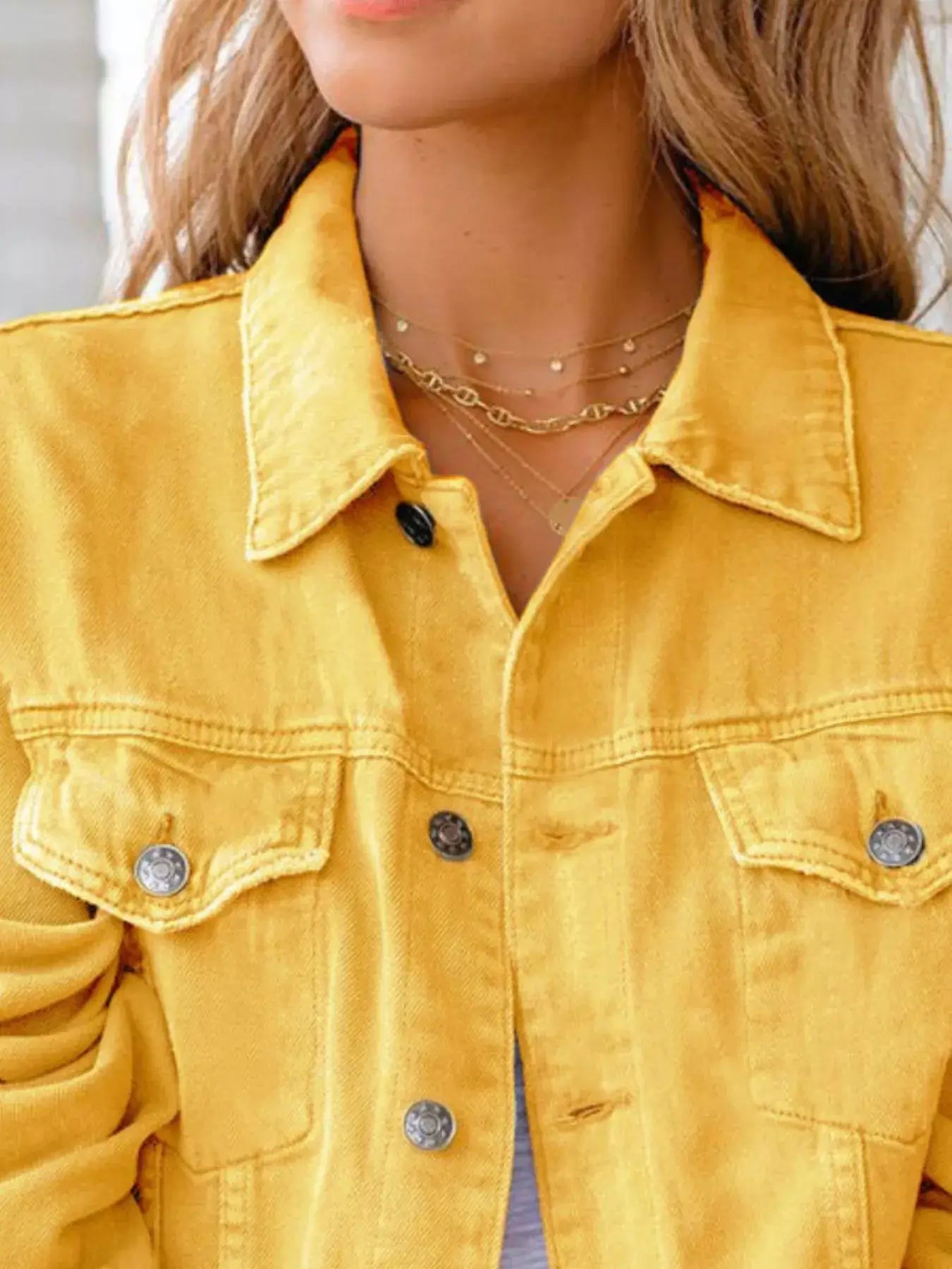 Close-up of a woman wearing a yellow button-up denim jacket with layered necklaces and a soft smile.
