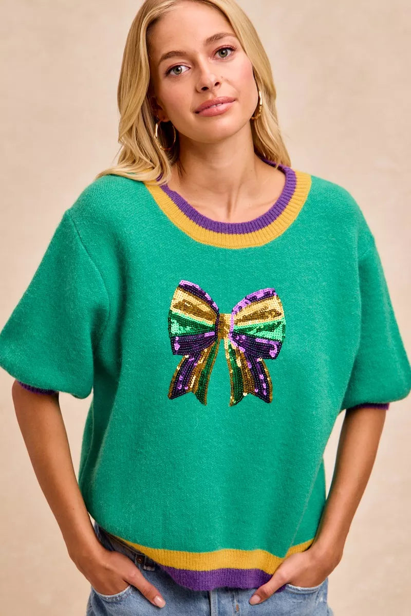 Woman wearing BiBi Mardi Gras Sequin Ribbon Patched Sweater featuring colorful sequin bow design.