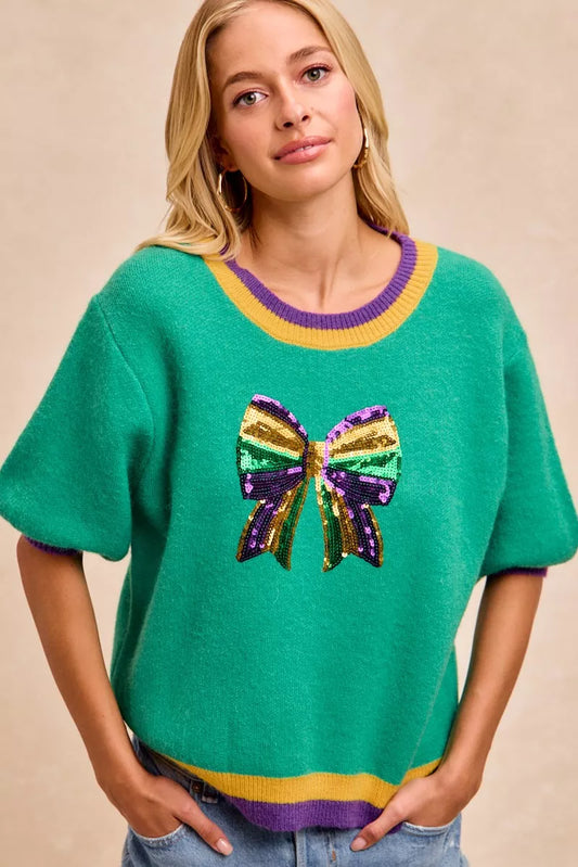 Woman wearing BiBi Mardi Gras Sequin Ribbon Patched Sweater featuring colorful sequin bow design.