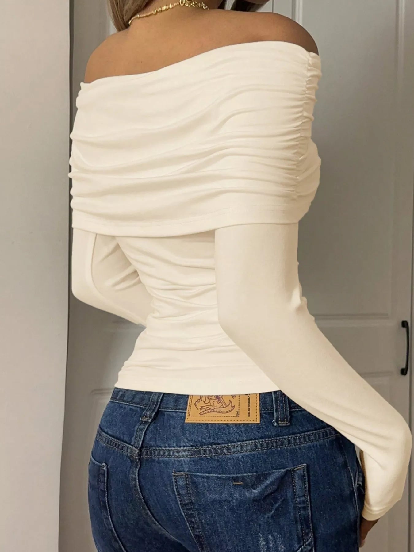 Back view of a model wearing an Off-Shoulder Ruched Long Sleeve T-Shirt in cream color, paired with denim jeans.