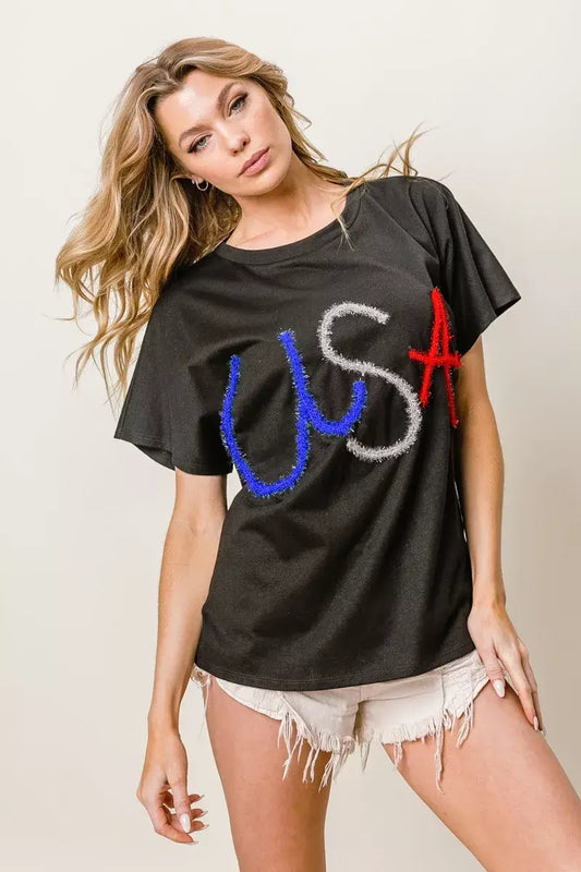Model wearing BiBi Metallic Letter Short Dolman Sleeve Top with 'USA' design in red, white, and blue letters, paired with denim shorts.