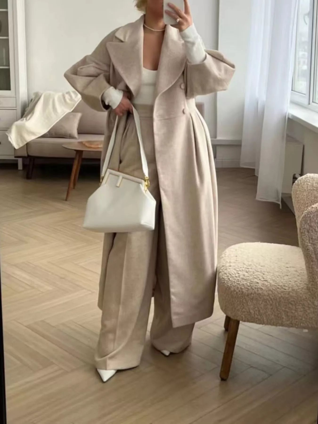 Elegant woman wearing a Double-Breasted Longline Coat with a tailored silhouette, holding a white bag, in a stylish indoor setting.